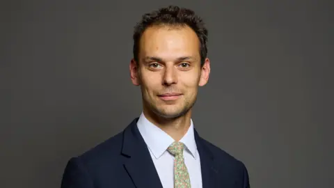 Josh Simons has dark hair, a pale green tie, white shirt and blue suit