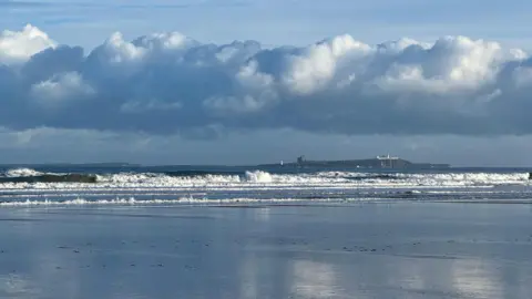 BBC WEATHER WATCHERS/ LIZ WILSON A chilly morning on Bamburgh beach with waves crashing onto the wet sand. A thick bank of cloud hangs over the beach.