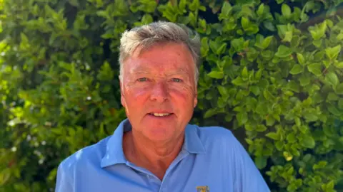 Russ Legg A photo of Russ Legg from the Tait's Hill Road Action Group. He is wearing a blue top and is standing in front of a green bush.
