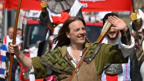 PA Media A man with long hair smiling and waving. He wears green and holds a bow