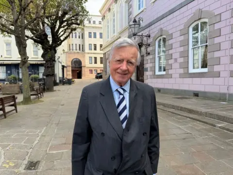 Deputy Sir Philip Bailhache is pictured outside the States Assembly. He is wearing a grey coat and a blue shirt. 