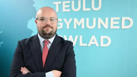 Reform UK Llŷr Powell in front of turquoise Reform UK branding with the Welsh words  Teulu, Gymuned, Gwlad meaning family, community, country. He has a beard and moustache and is wearing glasses, a light blue shirt, red tie and dark suit and is folding his arms.