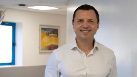 CUH NHS Foundation Trust Dr Lavinio smiles at the camera while standing in a corridor. He wears a white open necked shirt and is clean shaven with short dark hair. There is a colourful painting on the wall in the distance.