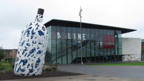 A general view of MIMA in Middlesbrough. The building is made of glass and has a flat roof. There is a large sculpture of a white and blue bottle to one side.