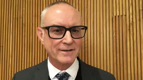 Jo Thewlis/BBC Fran Whymark, a man standing in front of a wall with wooden panels. He is wearing a suit with a white shirt and patterned tie and is looking directly at the camera.