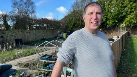 A man wearing a grey jumper stood in front of an allotment. He has short dark hair, swept to his right, and is clean shaven. He stands next to a wooden fence, behind which is a metal set of shelves for seed trays and a paved area leading to allotment beds.