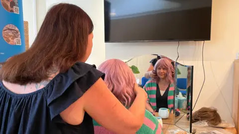 A woman with brown hair is fitting a pink wavy wig for a woman wearing a green and pink stripey cardigan.