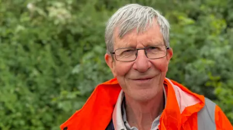 The head and shoulders of Harry Machin, who wears an orange high-vis jacket. He is looking at the camera smiling slightly. He has grey hair and black-framed glasses. Green foliage is behind him.