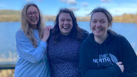 Three women lean against each other, each is laughing as they look at the camera. Behind them is a lake