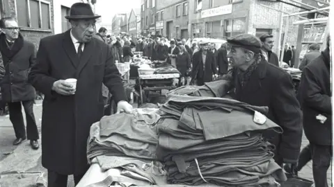 Black and white image of two men in hats and overcoats at an outdoor market. They are chatting over a pile of trousers on a market stall.