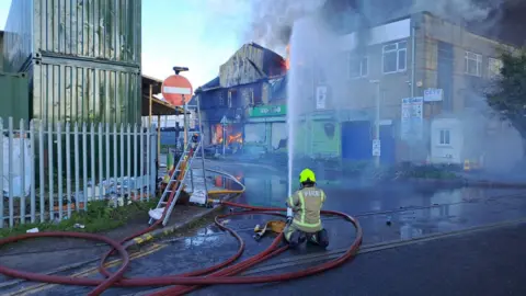 A firefighter kneeling in the road pointing hose spray up towards burning shell of a building 