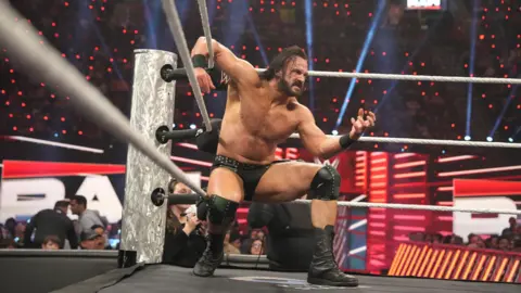 WWE Drew McIntyre in the ring, crouching by the turnbuckles, with one arm resting on the ropes. His face is in a grimace, while he is wearing black trunks and boots. He appears about ready to pounce on an opponent. 