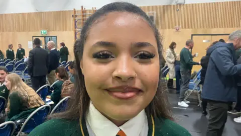 A teenage girl and long brown hair. She is smiling at the camera. She is standing in a hall with a group of people in the background.