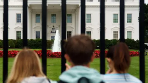 Three people are standing outside the White House behind black railings. One has dark hair in a ponytail, the other blonde long blond hair and the person in the foreground has short brown hair