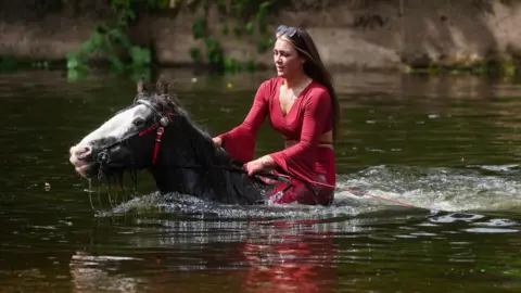 PA Media A woman rides the horse which is submerged in the River Eden up to its neck. The woman is wearing a red top and has long dark hair.