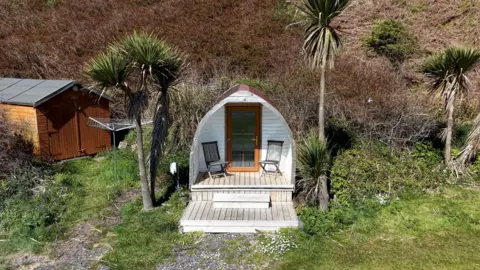 Galbraith A small pod building with decking and chairs in front of it sits beneath a couple of trees