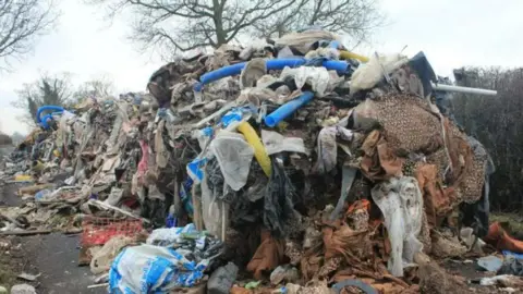 A huge pile of rubbish is strewn along a country road, with blue plastic tubes sticking out of it, among countless plastic bags that are bursting.
