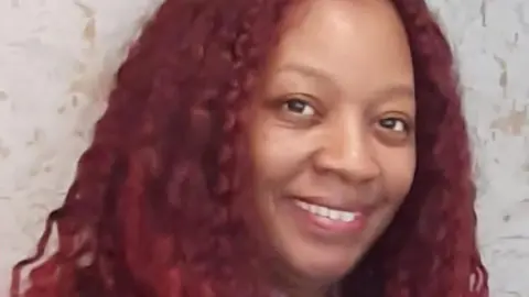 Faye Delaney has dyed, long dark red hair and is wearing a grey top under a black top. She is smiling at the camera.