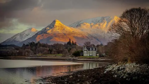 The sun shines on a mountain, with a loch in the foreground and a house in the centre