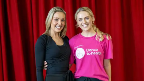 Arts Council of Wales The two women stood in front of a red curtain with their arms round each other. Amy on the left is wearing all black and has shoulder-length hair with blonde highlights. Emma on the right wears a pink t-shirt with the words 'Dance to health' in white. She has tied-back blonde curly hair. They are both looking at the camera and smiling.