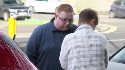 Daniel Wyke stood by a red car talking to another person stood in front of him. He has red hair, is wearing a dark jumper with rimless glasses on.