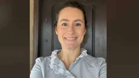 A woman smiling at the camera. She is wearing a blue and white striped top with white lace. Her brown curly hair is tied back. Behind her is a brown background. 