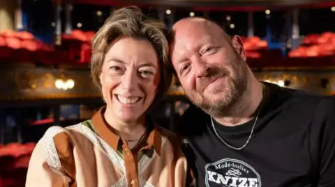 Nicole Taylor, with short highlighted brown hair and wearing a brown and peach shirt, and John Tiffany, a bald man with a short beard wearing a black t-shirt with a Knize logo, sit together smiling at the Lyceum Theatre in Edinburgh.