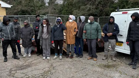 A group of people standing in front of a fence and a white van. They are wearing winter coats and not all of them are looking at the camera.