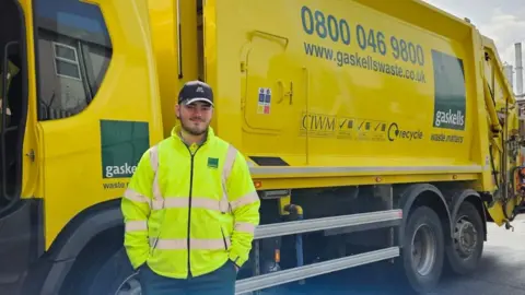 Gaskells Daniel Kinsella had a dark beard and is wearing a black baseball cap and yellow high-vis jacket. He is standing in front of a yellow bin lorry.
