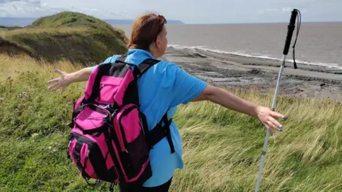 Becky Harrison Ms Harrison pictured from behind looking out over a coastal view. There is a muddy, sandy beach in front with sea and she stands on long grass looking over it. She has her arms outstretched and a pink backpack on.
