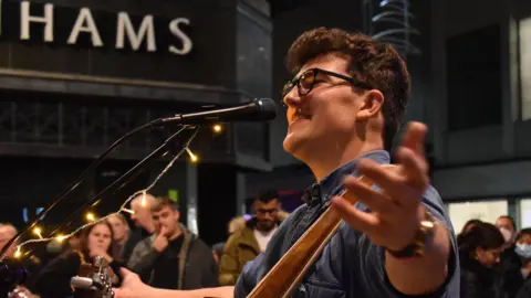A man with short brown hair and black-rimmed glasses stands with his arms wide open. He is talking or singing into a microphone and has a guitar around his neck.