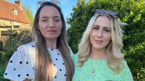 Richard Knights/BBC Ashleigh and April stand next to each other in a garden. They are both looking and smiling at the camera. Ashleigh has long brown hair with glasses on top of her head, and she is wearing a white blouse with black dots. April has shoulder length blonde hair with glasses on her head. She is wearing a green top with flowers on it and a necklace around her neck.