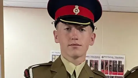 Jack Burnell-Williams in ceremonial uniform and peaked cap stands indoors, facing the camera, with training posters visible on the wall behind him.