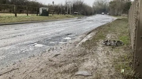 BBC A main road, which is pockmarked with ridges and craters. There is some debris on the side of the road, including a hubcap. It is a wet day so the holes are puddles.