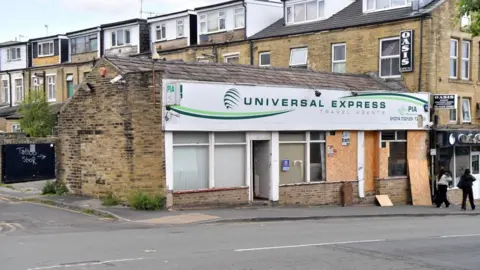 LDRS A single storey brick travel agents building. Some windows are boarded up and there is a memorial stone outside.