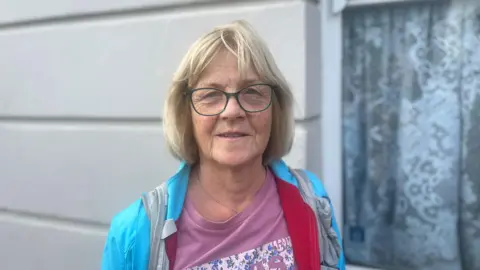 a blonde woman wearing a blue coat with pink top, she is wearing glasses.