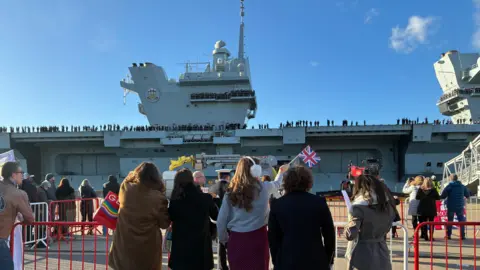 A group of women stands with their backs to the camera, waving their hands or Union Jack flags at HMS Prince of Wales docks at Portsmouth Naval Base.