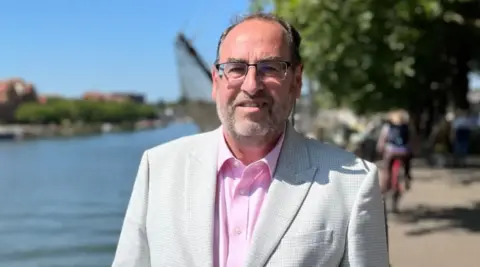 Mr Dyer smiles into the camera. He's wearing a white suit jacket with a pink shirt and has glasses. Behind him, the Bristol harbour is seen in the sunshine. It is blurred. 