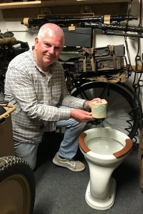 Graeme Delanoe kneels holding a roll of occupation-era toilet roll above a toilet bowl with is white porcelain with two brown wood panels around the top of the bowl. Behind are various WW2 items stacked on shelves.