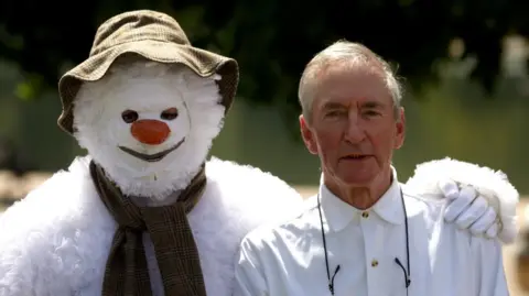 PA Media Author Raymond Briggs stood next to a person dressed as a snowman. The snowman, left, has its arm around Mr Briggs.