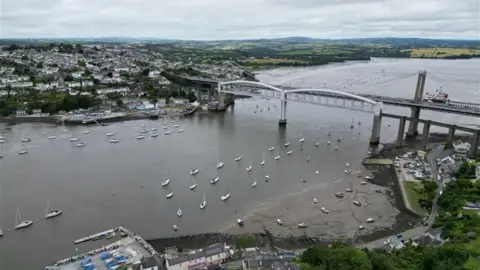 A drone shot of Tamar Bridge. The train bridge and car bridge are visible. There are sailing boats on the river Tamar. Saltash is on the left and Plymouth is on the right. There are pastel houses on both sides. The water is a dark grey. The sky is full of clouds. 