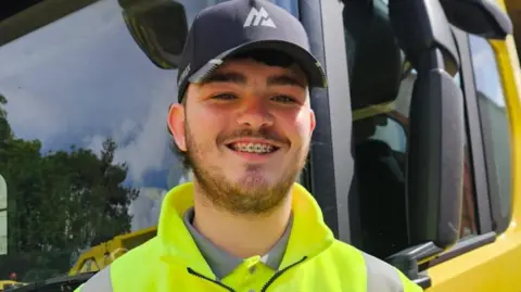 Daniel Kinsella has a dark beard and is wearing a black baseball cap and yellow high-vis jacket. He is standing in front of a yellow bin lorry and smiling for the camera.
