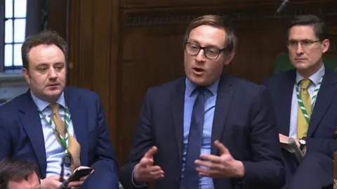 parliamentlive.tv Olly Glover gives a speech in parliament. He is wearing a blue shirt, blue tie and blue suit and is surrounded by other MPs.