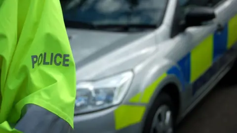 Police officer's arm in a hi-vis jacket with a police car behind 