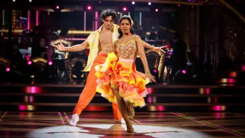 Julian Caillon and Balvinder Sopal dance in yellow and orange dance costumes on stage in Strictly. Caillon has his arms out standing behind Sopal, who is stepping forward