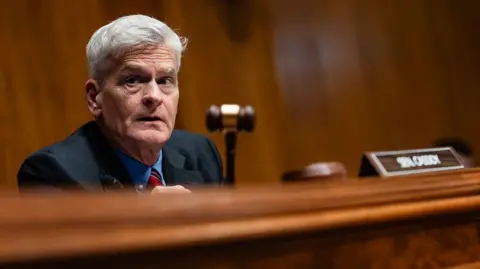 Getty Images Bill Cassidy, who has grey hair and a blue shirt with a red tie, holds a gavel during a hearing on Capitol Hill.