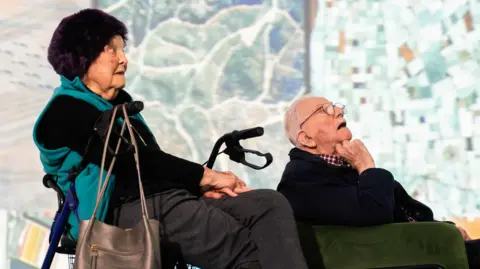 This image shows two older adults sitting side by side indoors, positioned in front of a large display that looks like an aerial map or satellite view.
In the foreground, one person is seated on the left using a wheeled walker. The walker has a metal frame and a small storage basket. This person is wearing dark clothing with a teal or green layer draped over the shoulders and has a handbag resting on the walker.