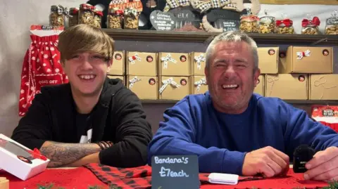 Two men standing and smiling side by side. The man on the left is younger, with long brown hair and a tattoo on his arm. The man on the right is laughing, wearing a blue sweatshirt and short grey hair. There are boxes piled behind them. 