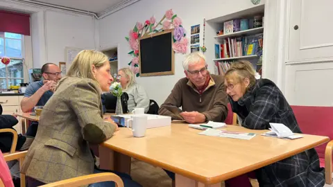 Wera Hobhouse and an older couple sitting opposite each other on a table on pink chairs with a few bookshelves in the background in what looks like a community hall or room.