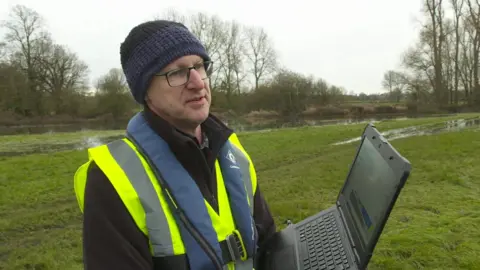 A picture of Nick Everard, who has a hi-vis jacket on and a black and blue woolly hat. He is holding a laptop computer in a waterlogged field.